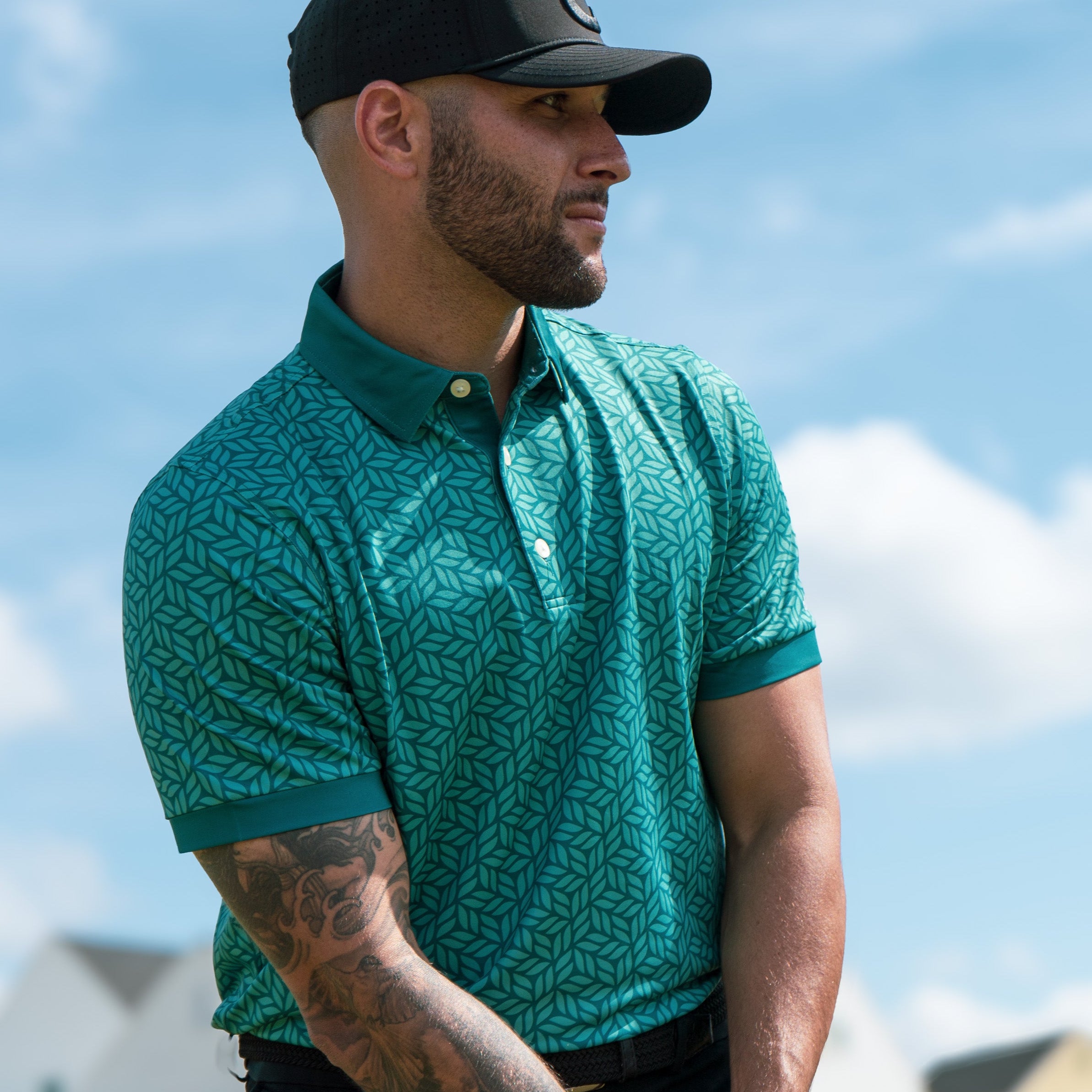 Man wearing a green patterned polo shirt and black cap outdoors with houses and blue sky in the background