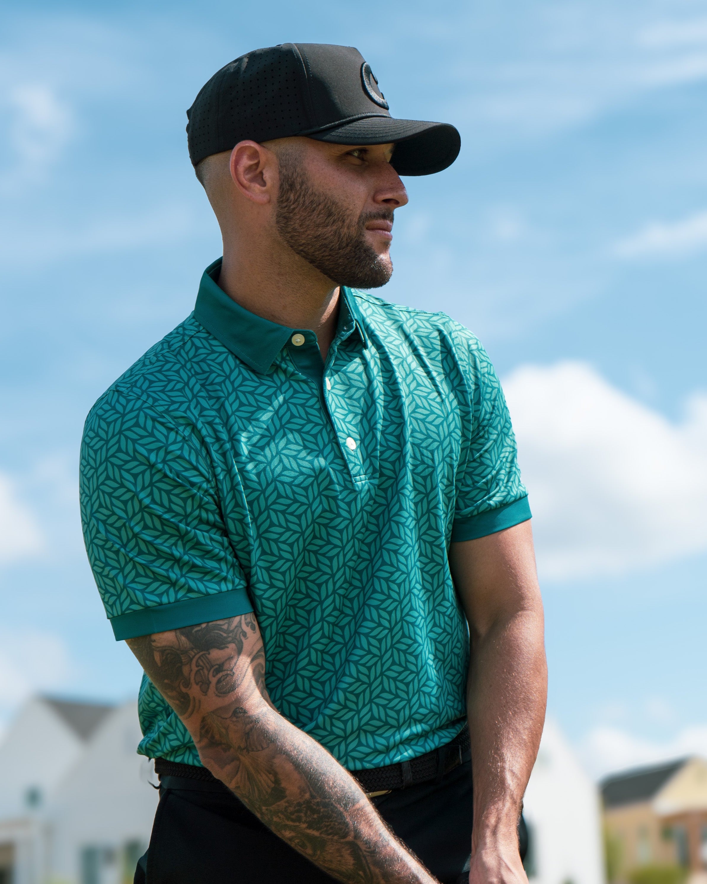Man wearing a green patterned polo shirt and black cap outdoors with houses and blue sky in the background